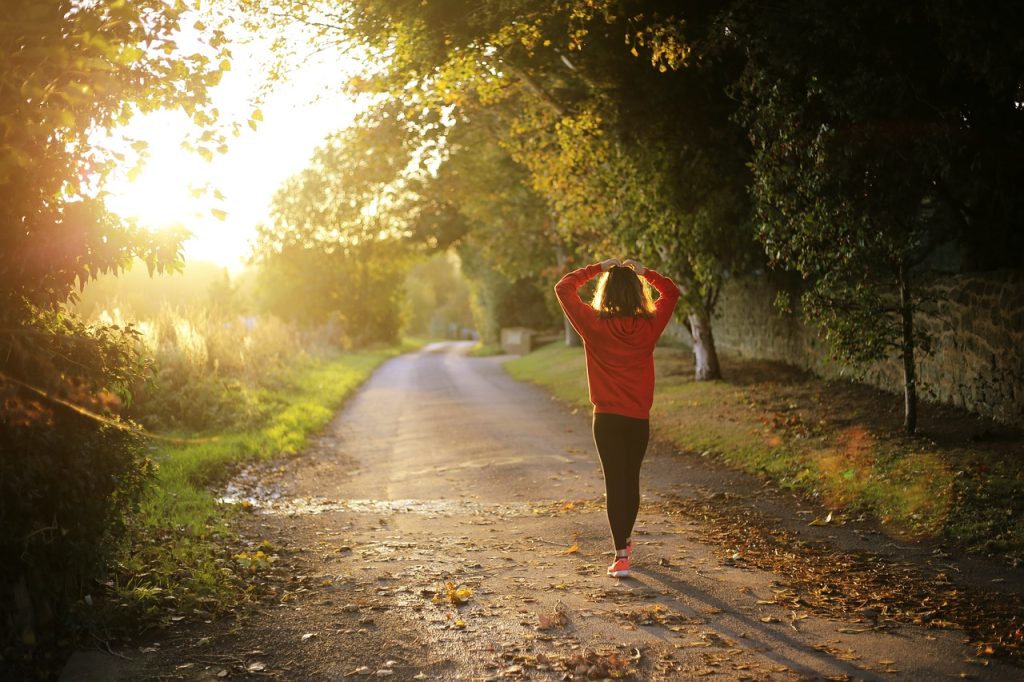 This image shows the backside of a woman walking on dirt path somewhere who is wearing a bright orange sweater or sweatshirt and black leggings, plus pink sneakers and who has her heads toughing the top of her head. It is fall day and the sun is shining thru the branches on trees and thru leaves, but some leaves have already fallen and cover the path she is walking on.
