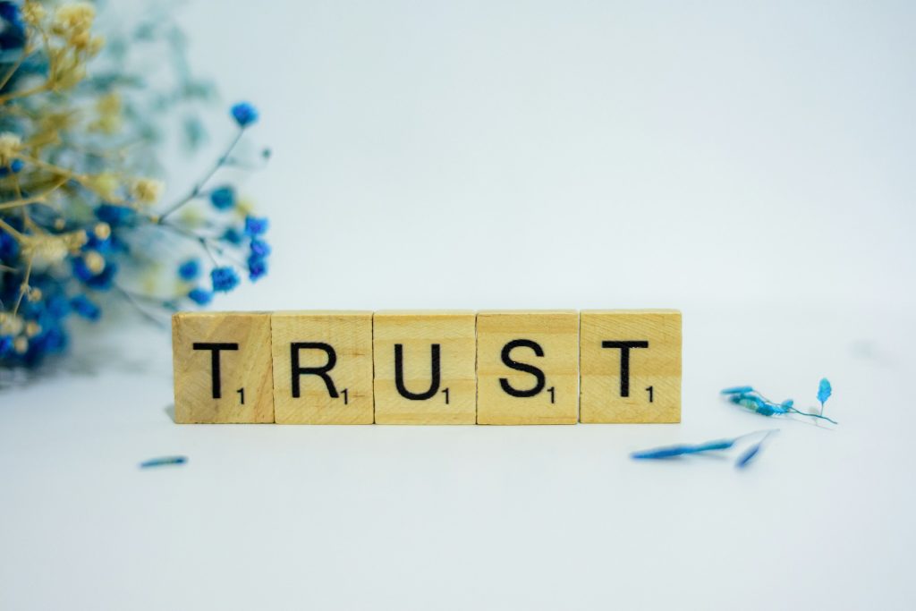 This is the rectangular image of Scrabble game letters standing up and spelling "Trust" and includes some blue flowers surrounding the letters that appear on a white background.