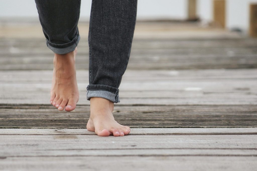 this image shows the legs of woman below the knees who is walking on wooden pier and who is wearing jeans and who is bare-footed.
